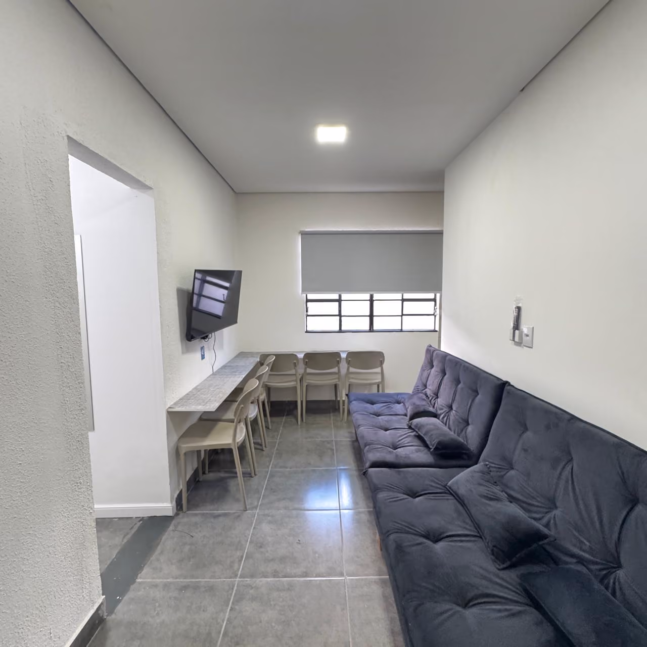 Quarto moderno com cama branca, almofadas bege, cobertor, cadeira de escritório branca e três quadros decorativos abstratos na parede, com janela mostrando prédios ao fundo.