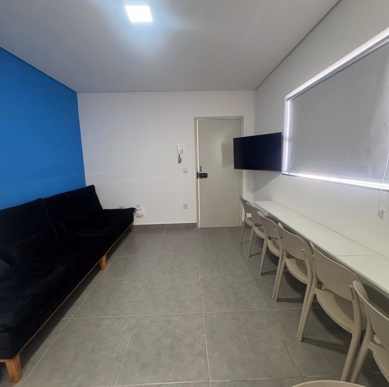 Quarto moderno com cama de solteiro branca com cobertor bege e roupas dobradas, TV preta sobre bancada branca, cadeira branca de escritório e duas pinturas abstratas com folhas e formas geométricas em tons terrosos na parede cinza clara.